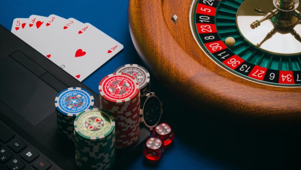 A close-up of poker chips, cards, and a roulette wheel next to a laptop and dice